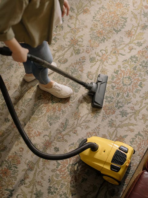 A person wearing beige sneakers and blue jeans is using a vacuum cleaner with a yellow body and black hose to deep clean a patterned area rug featuring floral motifs in muted tones of beige, green, an