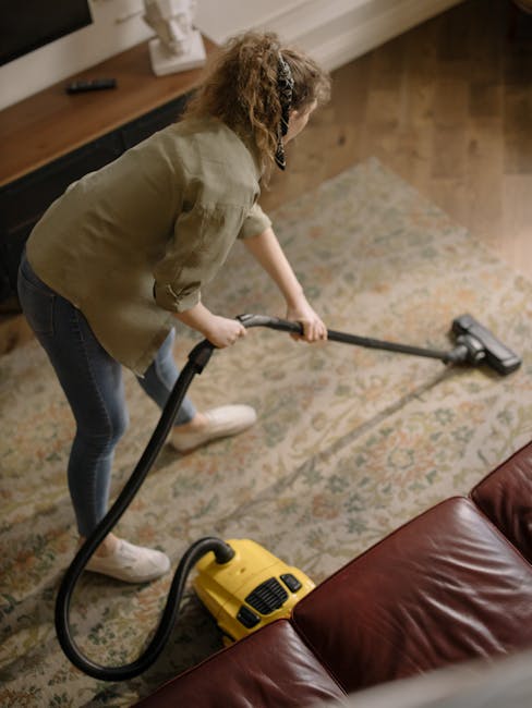A person wearing beige sneakers and blue jeans is using a vacuum cleaner with a yellow body and black hose to deep clean a patterned area rug featuring floral motifs in muted tones of beige, green, an