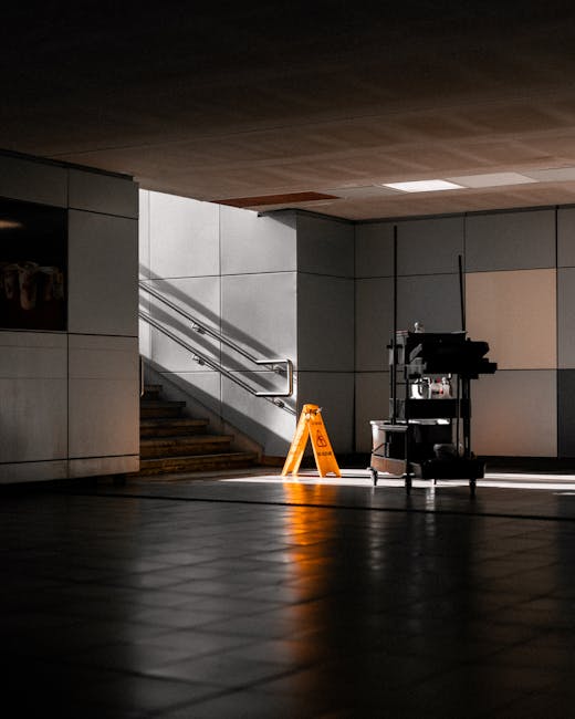 A dimly lit underground commercial space with dark tiled flooring and beige wall panels. Sunlight streams in, casting shadows across the floor and illuminating a bright yellow cleaning caution sign pl